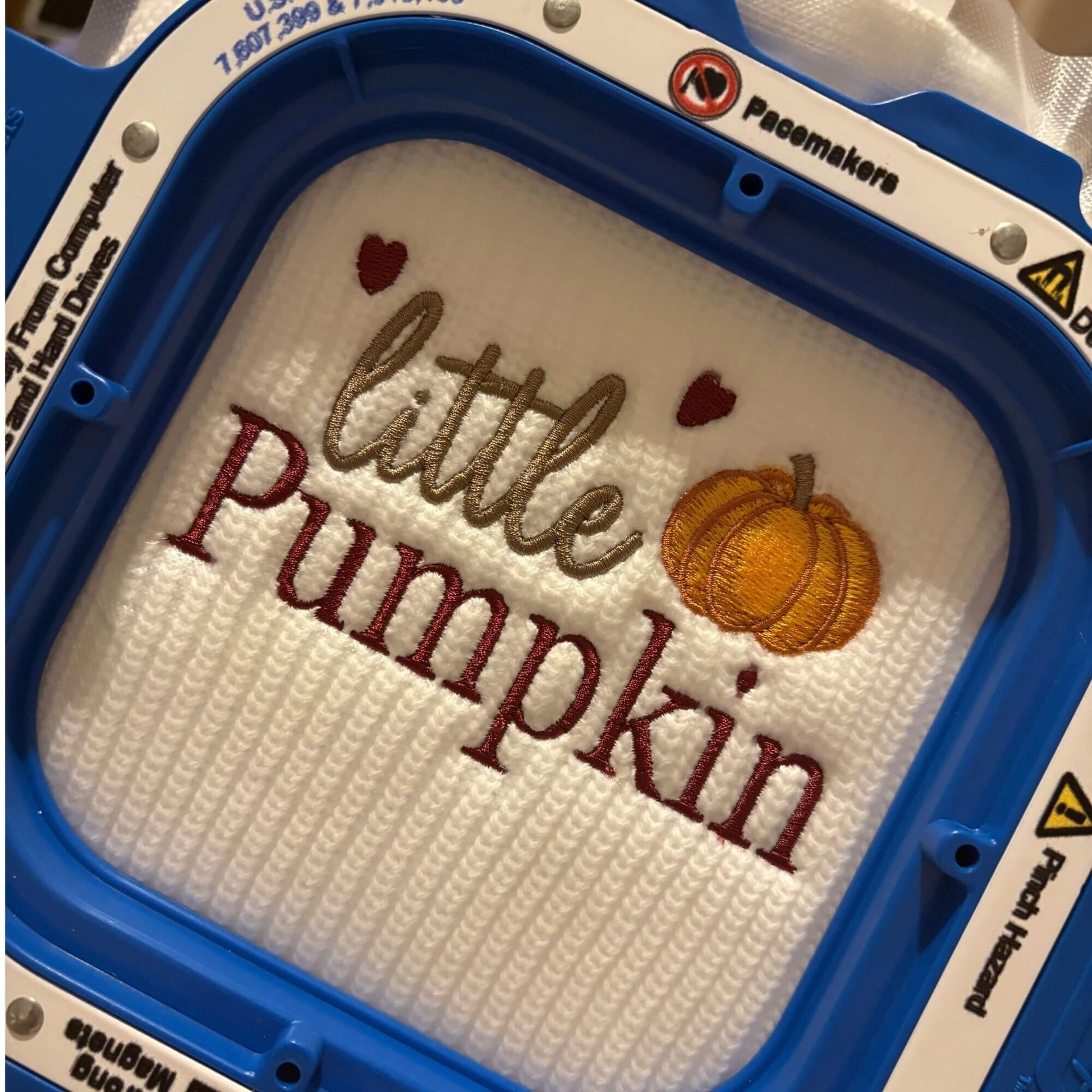 A close-up of a completed embroidery design inside a blue mighty hoop, featuring the words 'little Pumpkin' in brown and burgundy thread with two small hearts and an orange pumpkin motif on white  white baby cardigan.