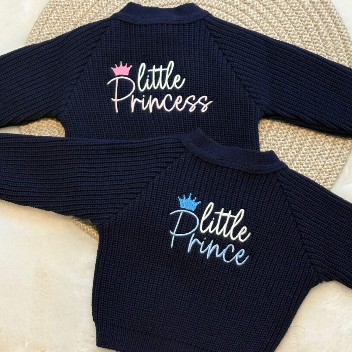Two navy blue ribbed knit baby cardigans laid flat on a woven mat. The embroidery reads 'little Princess' in white script with a pink crown, and the  'little Prince' in white script with a blue crown.