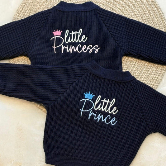 Two navy blue ribbed knit baby cardigans laid flat on a woven mat. The embroidery reads 'little Princess' in white script with a pink crown, and the  'little Prince' in white script with a blue crown.