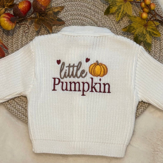 A white ribbed knit baby cardigan sweater with 'little Pumpkin' embroidered on the back, styled on a textured beige blanket with autumn leaves and pumpkins.