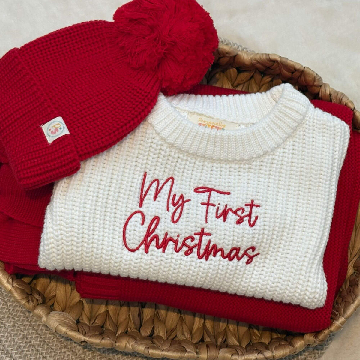 A folded white knit romper with red embroidered with 'My First Christmas'. paired with matching pompom bobble hat and blanket folded in a basket