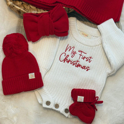 A baby holiday knit set laid flat on a white faux-fur surface, including a white long-sleeve knitted bodysuit with red embroidered text reading 'My First Christmas,' a red bow headband, a red pom-pom beanie, and red booties. A woven basket and folded red knit blanket are partially visible in the background.