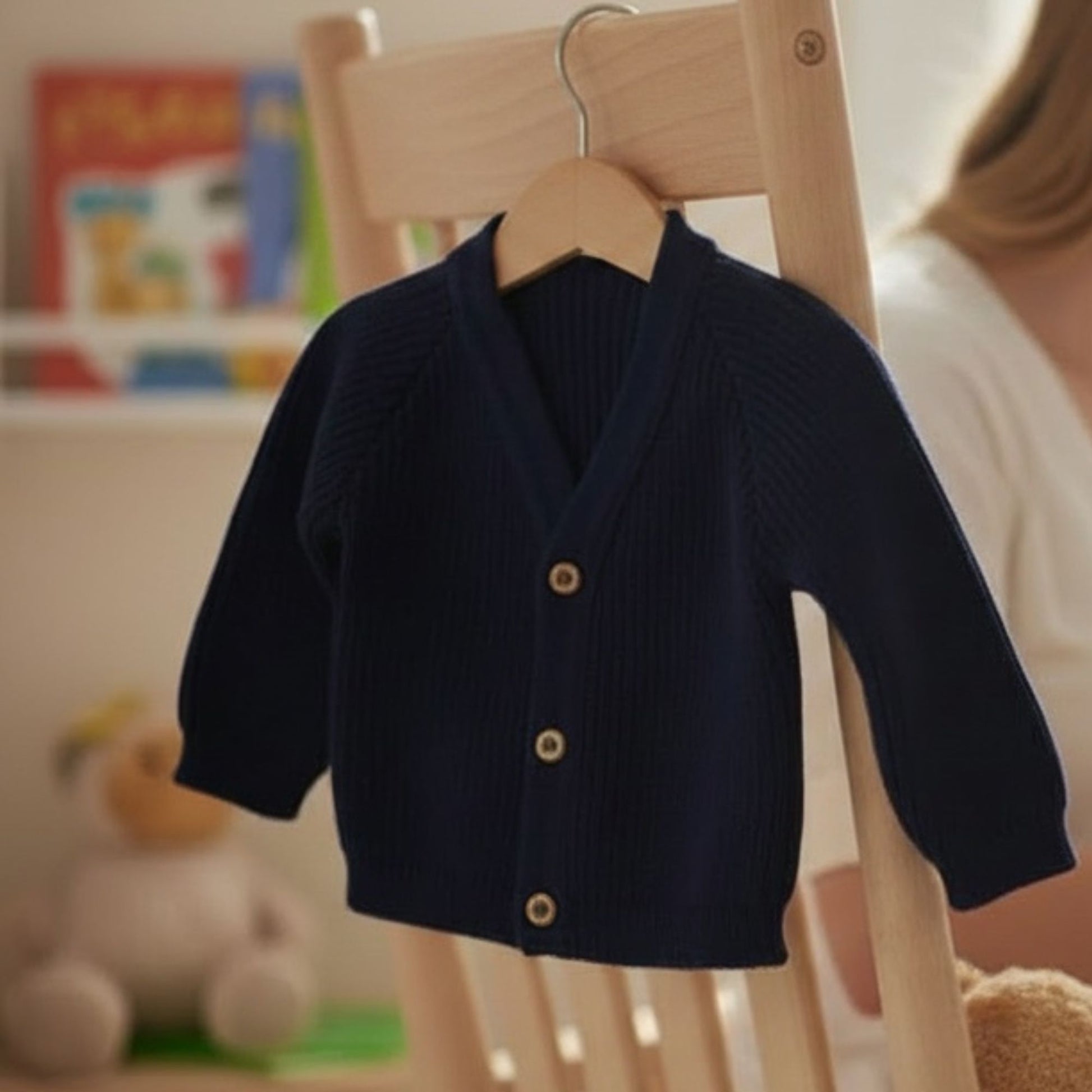 A navy blue dandelion ribbed knit baby cardigan with three wooden buttons, hanging on a wooden hanger. Personalised with babies name using embroidery 