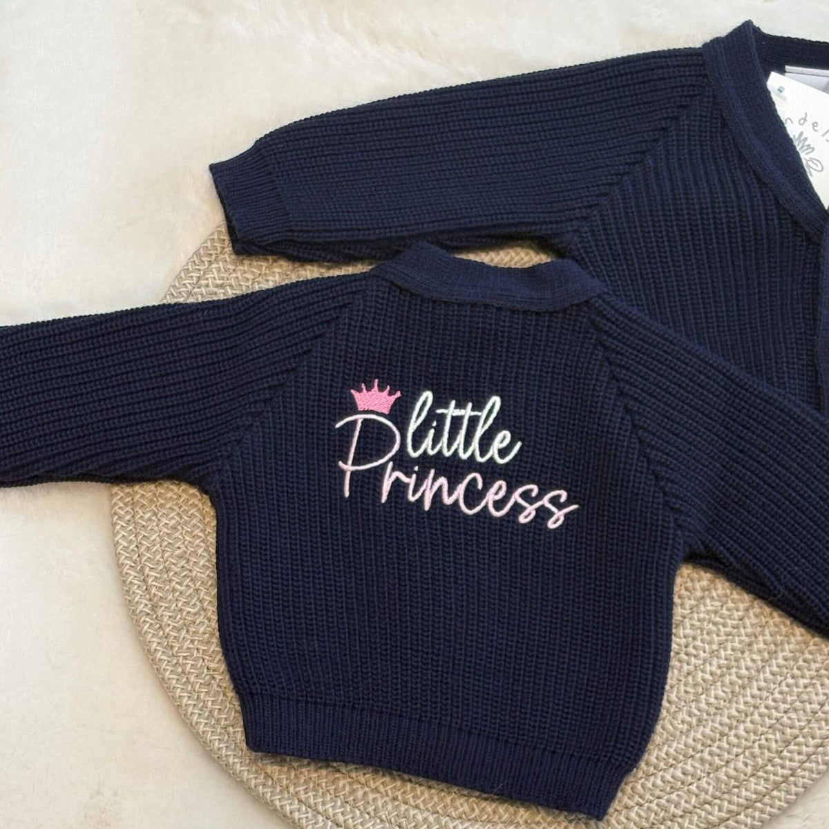A navy blue ribbed knit baby cardigan with 'Little Princess' embroidered on the back in white and pink, styled on a woven round mat.