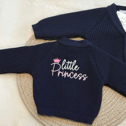 A navy blue ribbed knit baby cardigan with 'Little Princess' embroidered on the back in white and pink, styled on a woven round mat.