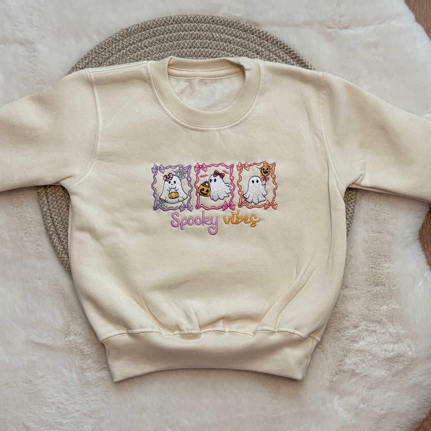 An oat colour crewneck sweatshirt featuring a 'Spooky Vibes' graphic with three cartoon ghosts in pastel frames, laid flat on a textured woven mat over a soft white faux fur surface.
