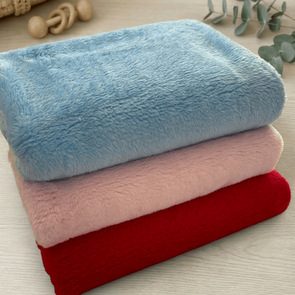 Three folded plush blankets in light blue, dusty pink, and deep red are stacked on a light wood surface, with a woven basket and eucalyptus leaves in the background.