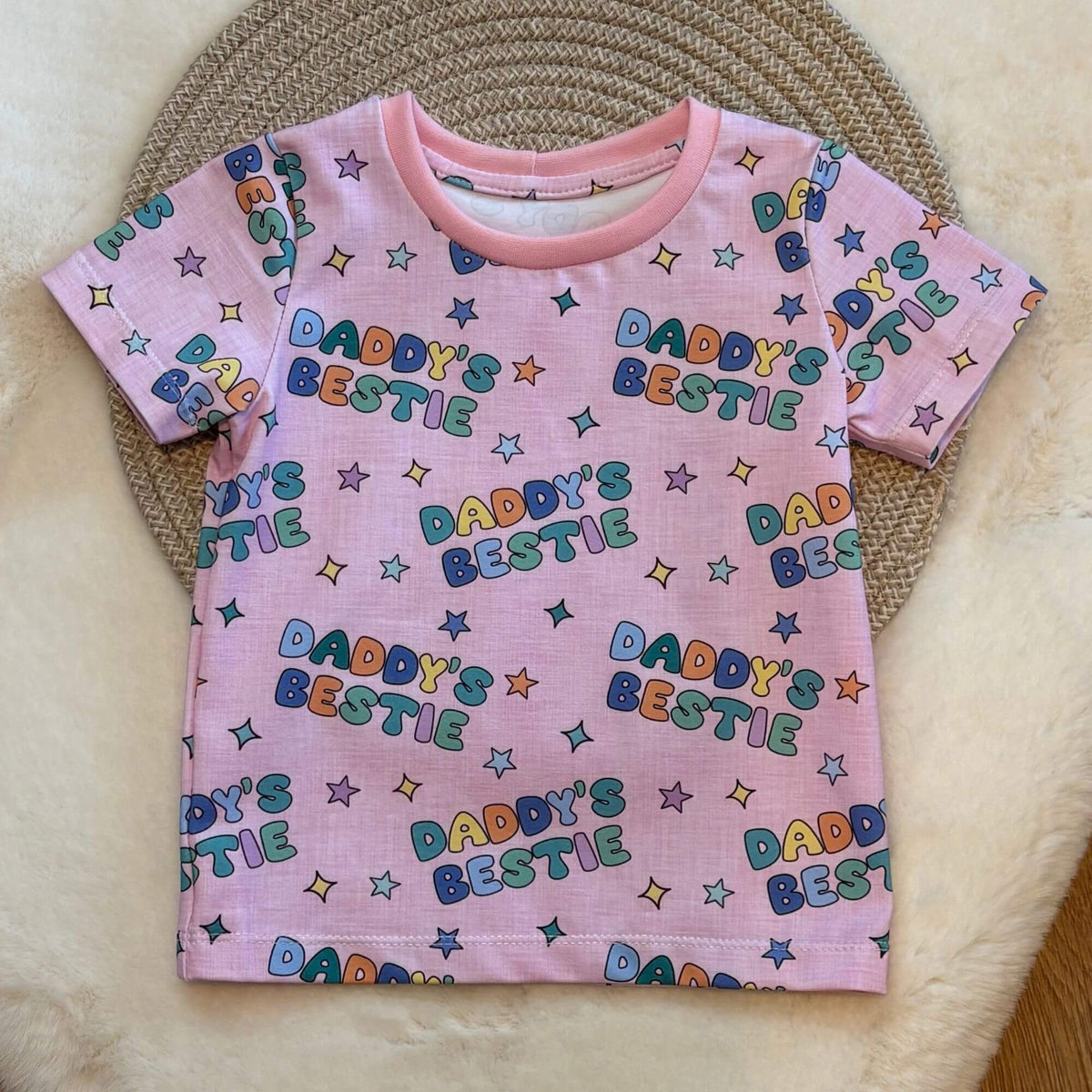 A pink short-sleeve baby t-shirt with an all-over 'Daddy's Bestie' text and star pattern, laid flat on a woven circular mat over a cream faux fur surface.