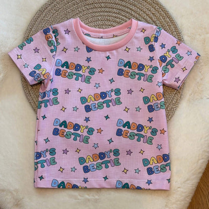 A pink short-sleeve baby t-shirt with an all-over 'Daddy's Bestie' text and star pattern, laid flat on a woven circular mat over a cream faux fur surface.