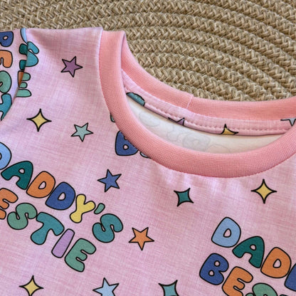 A close-up of a pink short-sleeve t-shirt with a colorful retro-style print reading 'Daddy's Bestie' in multicolored letters and stars, laid flat on a woven straw mat.