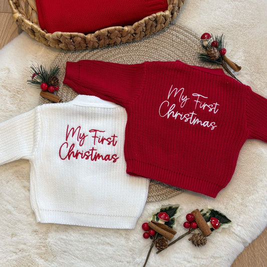 Two ribbed knit baby cardigans laid flat on a cream faux-fur surface, one white and one red, both embroidered with the phrase 'My First Christmas' in script. The scene includes holiday props such as pine sprigs, berries, cinnamon sticks, and a woven basket.