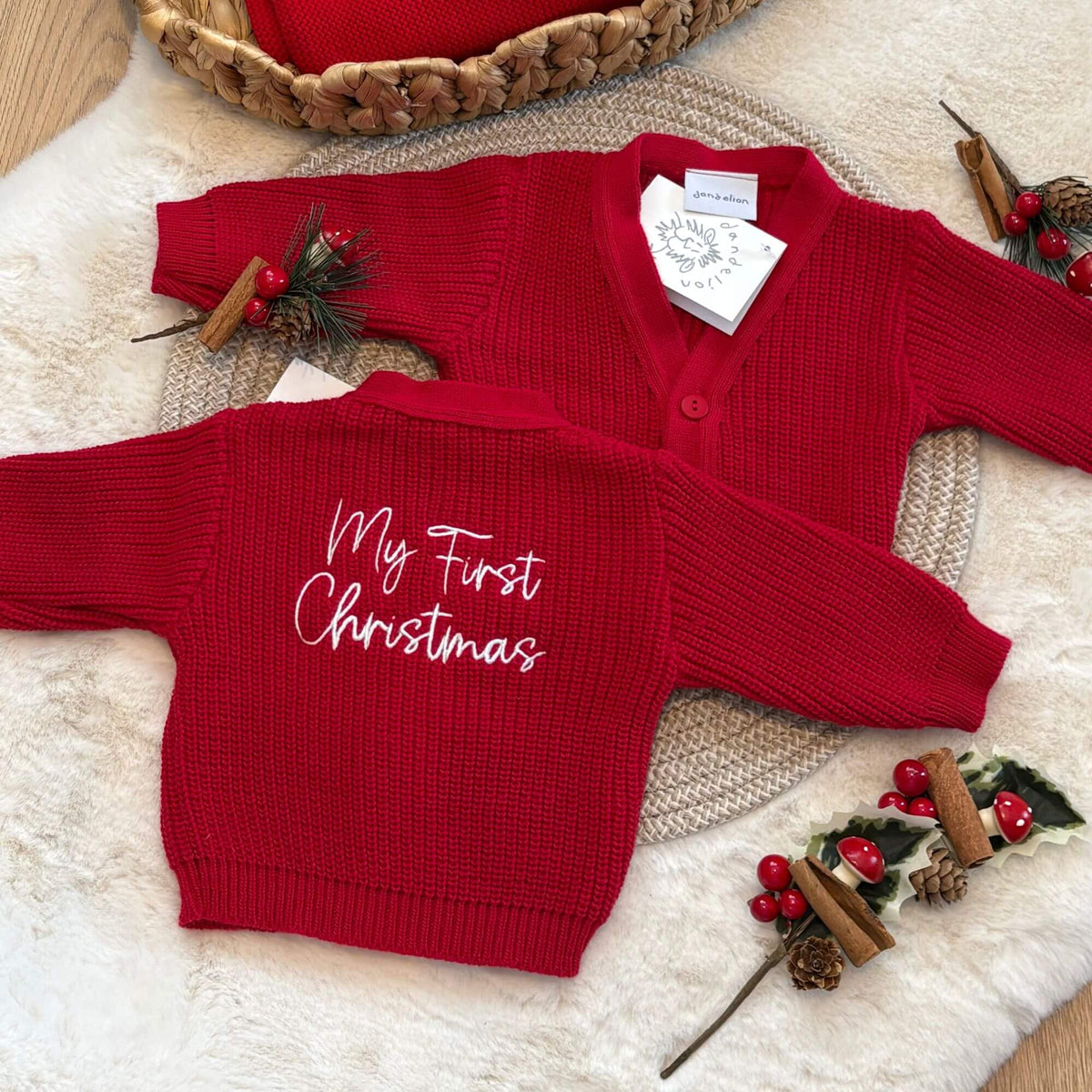 A red knit baby cardigan sweater with long sleeves and white script text reading 'My First Christmas' on the back shown on a cream faux-fur surface with holiday props. A second view of the same cardigan is placed behind it, with a white product tag visible.