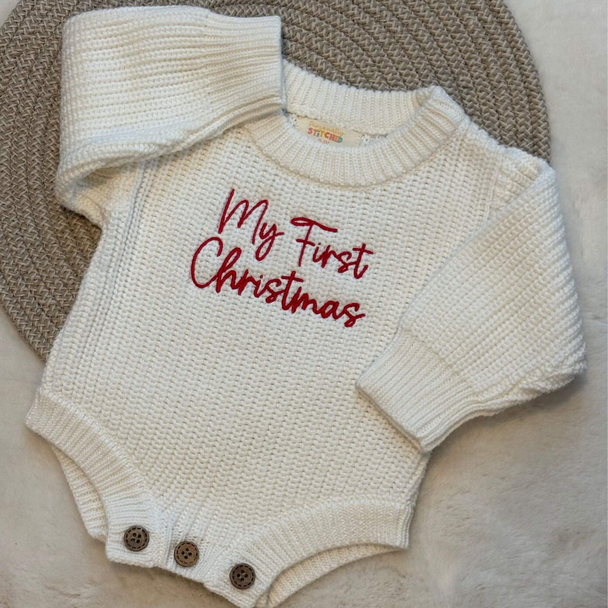 An off-white organic knit baby romper with 'My First Christmas' embroidered in red script across the chest, laid flat on a woven brown mat.