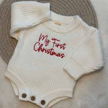 An off-white organic knit baby romper with 'My First Christmas' embroidered in red script across the chest, laid flat on a woven brown mat.