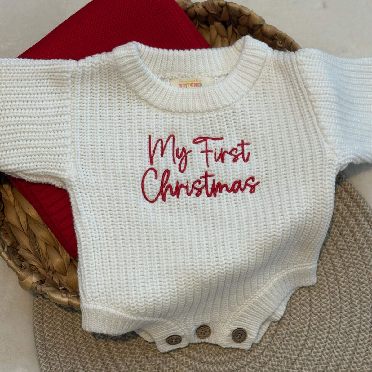 An off-white ribbed knit baby bodysuit with 'My First Christmas' embroidered in red script across the chest, displayed on a woven mat with a red knit personalised blanket underneath.