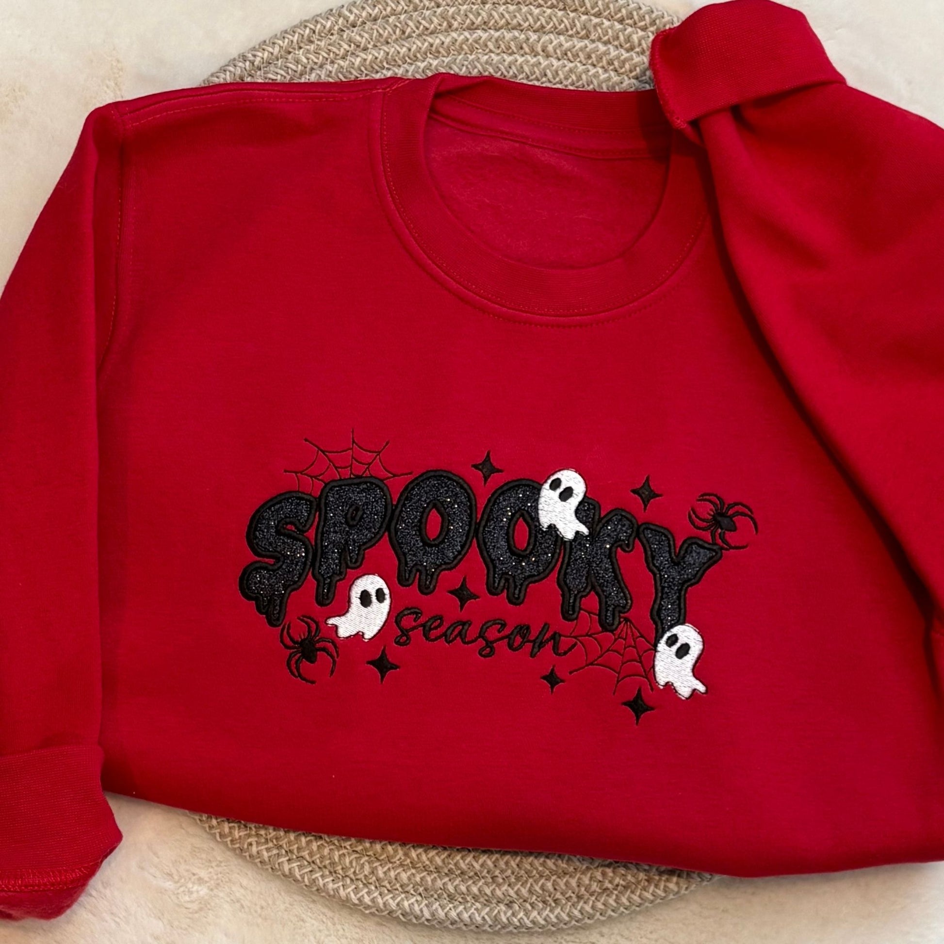 A red long-sleeve sweatshirt laid flat on a woven placemat, featuring a black glittery 'Spooky Season' graphic with white ghosts and black spider details.
