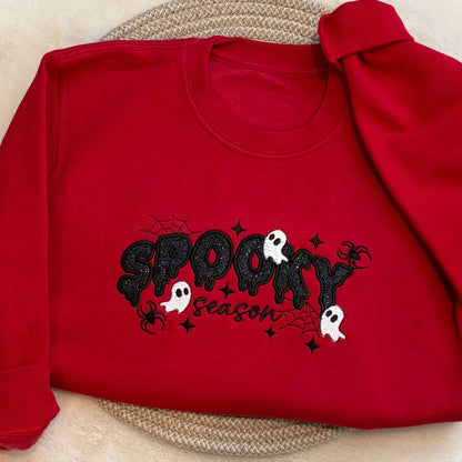 A red long-sleeve sweatshirt laid flat on a woven placemat, featuring a black glittery 'Spooky Season' graphic with white ghosts and black spider details.