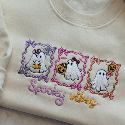 Close-up of an oat-colored sweatshirt featuring embroidered cartoon ghosts in purple, pink, and orange frames, with the text 'Spooky vibes' stitched below.