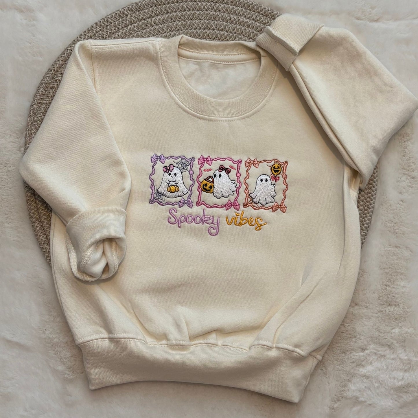 Oat coloured crewneck sweatshirt featuring a 'Spooky Vibes' graphic with three cartoon ghosts in purple, pink, and orange frames, laid flat on a textured woven mat.