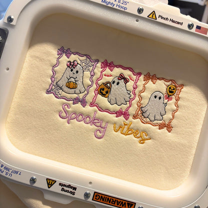 A close-up of a machine embroidery design featuring three cute cartoon ghosts in purple, pink, and orange frames, with the words 'Spooky vibes' stitched below in matching pastel colors. The embroidery is inside an embroidery hoop