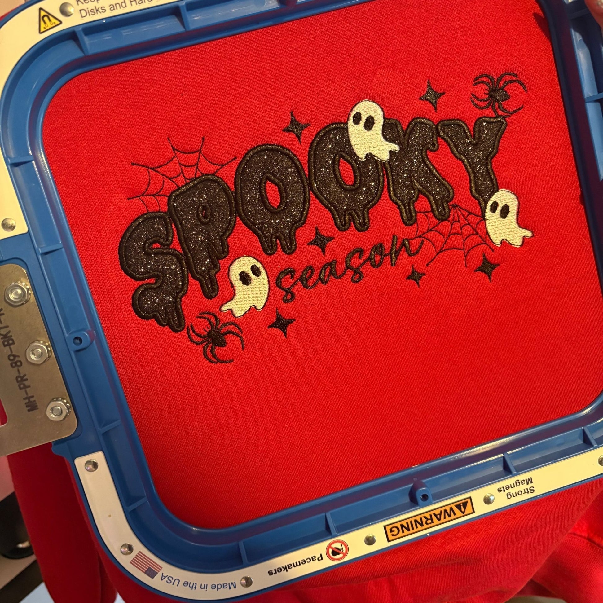 A close-up of a red fabric design inside a blue embroidery machine frame, featuring the phrase 'Spooky Season' in dark glittery letters with white ghosts, black spiderwebs, and black stars.