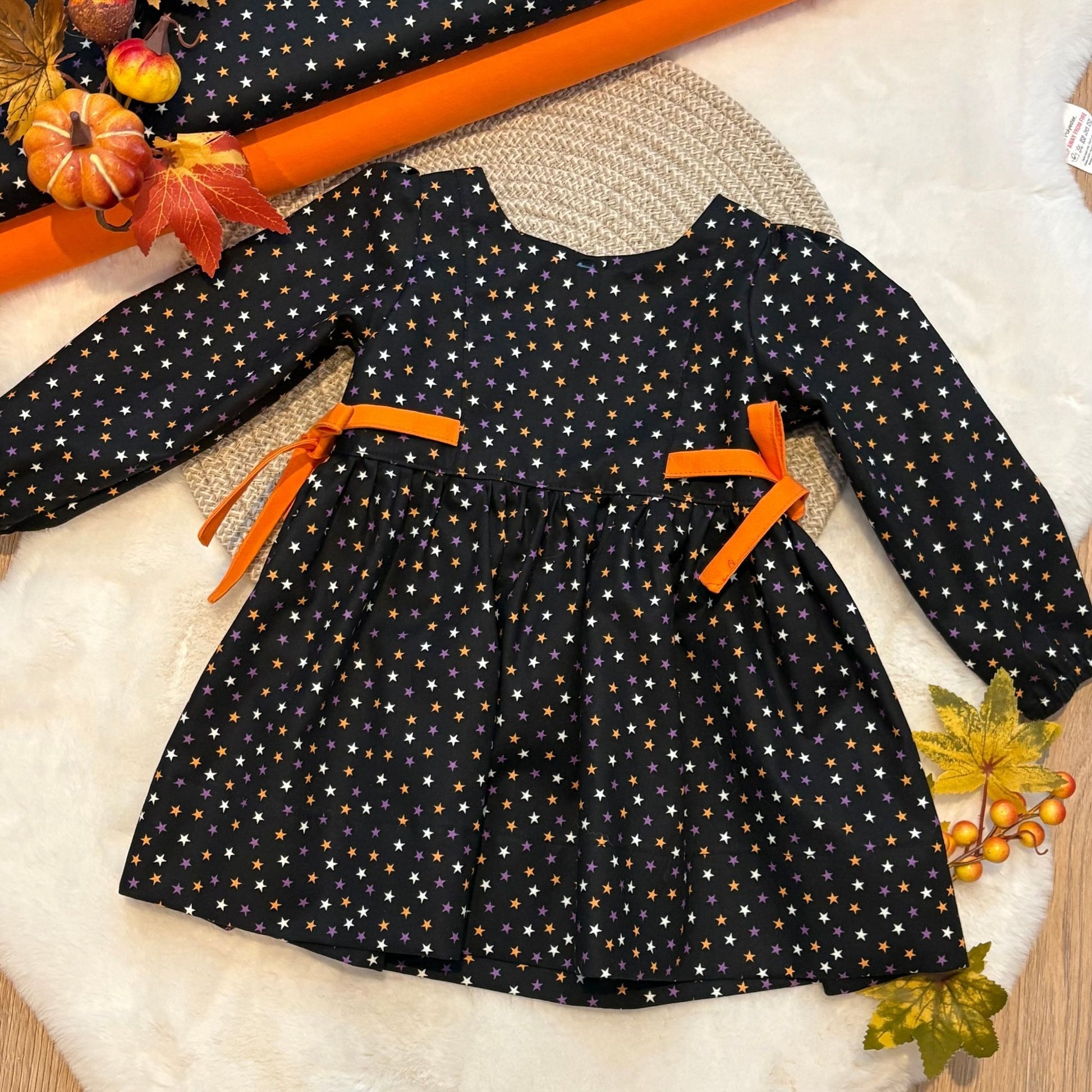 A long-sleeve baby dress with a black base, white and orange star pattern, and orange ribbon ties at the waist. 