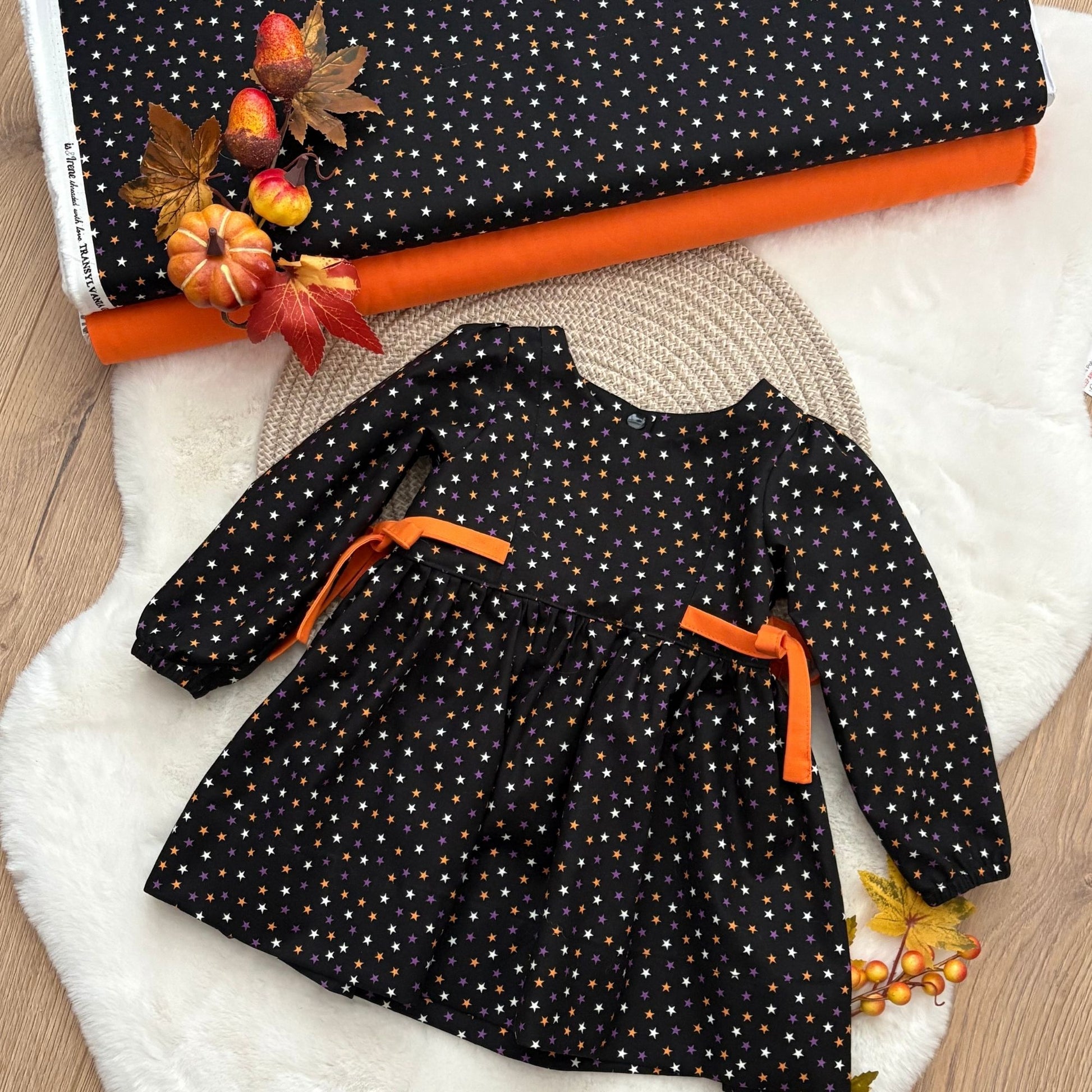 A toddler girl's long-sleeve dress with a black base, white and orange polka dots, and orange ribbon ties at the waist.