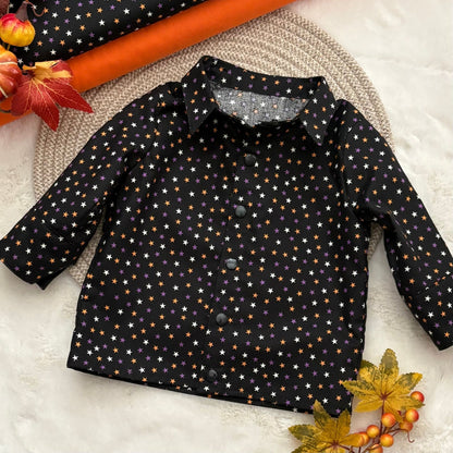A baby long-sleeve button-up shirt with a black base and an all-over pattern of small white, orange, and purple stars, styled in a flat lay with autumn leaves, a pumpkin, and a woven placemat.