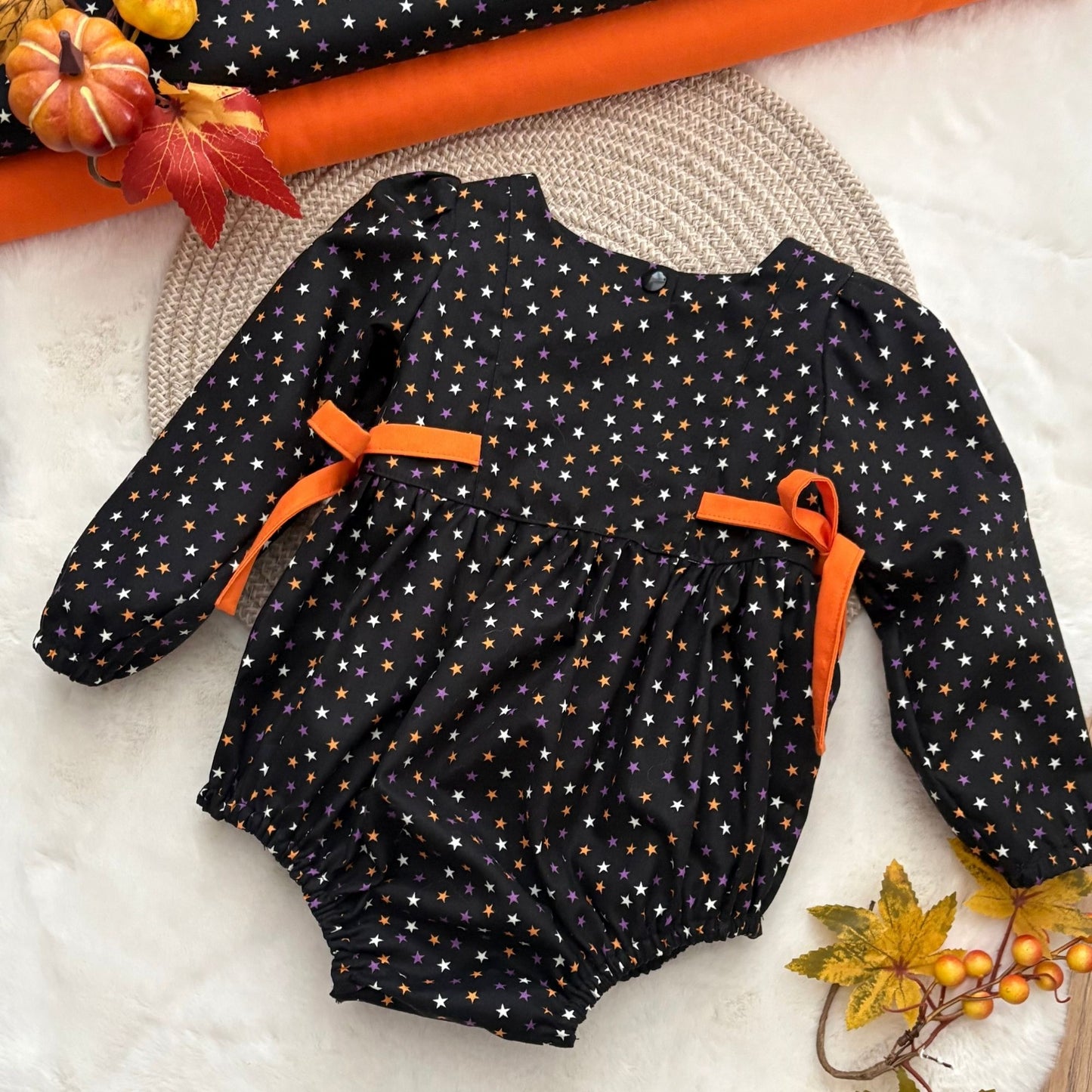 A baby long-sleeve romper in black with a white and orange star pattern, featuring orange ribbon bows at the waist