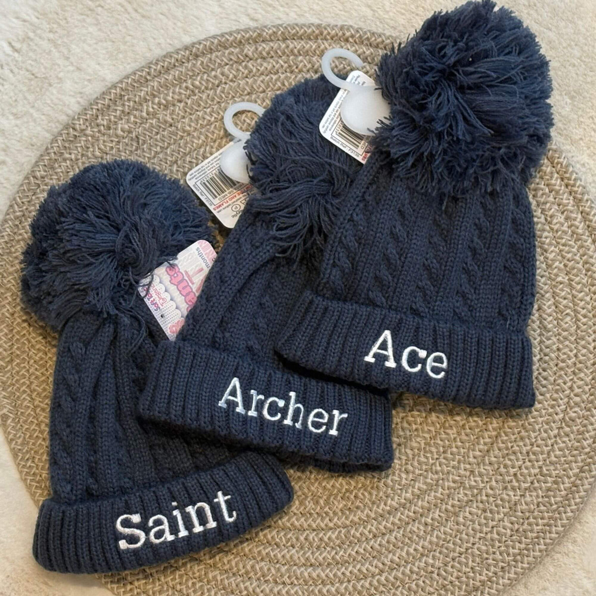 Three steel blue cable-knit baby beanies with large pom-poms are arranged on a woven round mat. The beanies are personalised with white embroidered name.
