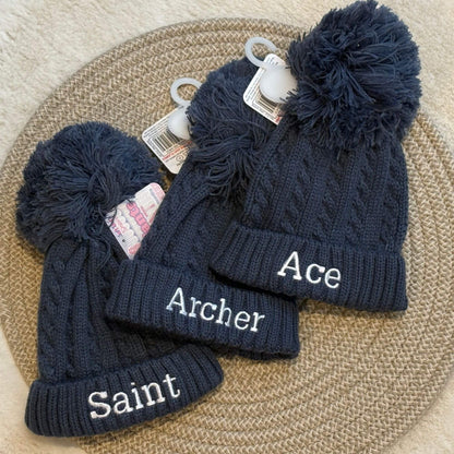 Three steel blue cable-knit baby beanies with large pom-poms are arranged on a woven round mat. The beanies are personalised with white embroidered name.