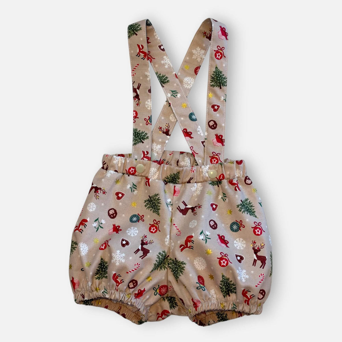 A pair of baby bloomers with adjustable suspenders, featuring a festive Christmas print with reindeer, trees, candy canes, and snowflakes on a light beige background.