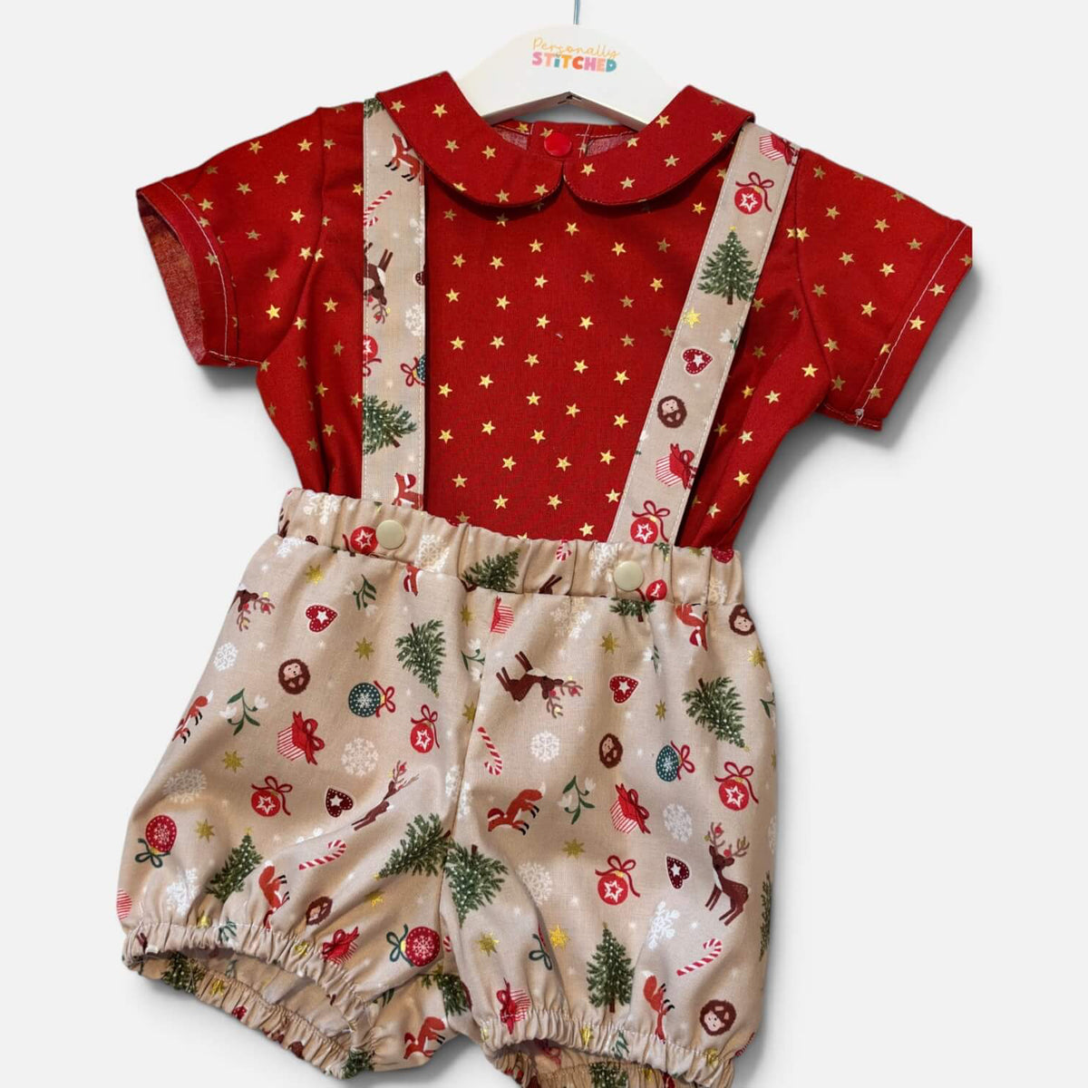 A baby romper set featuring a red short-sleeve top with gold stars and a Peter Pan collar, paired with beige bib-style shorts printed with Christmas motifs like trees, ornaments, and candy canes, shown on a white hanger against a light gray background.