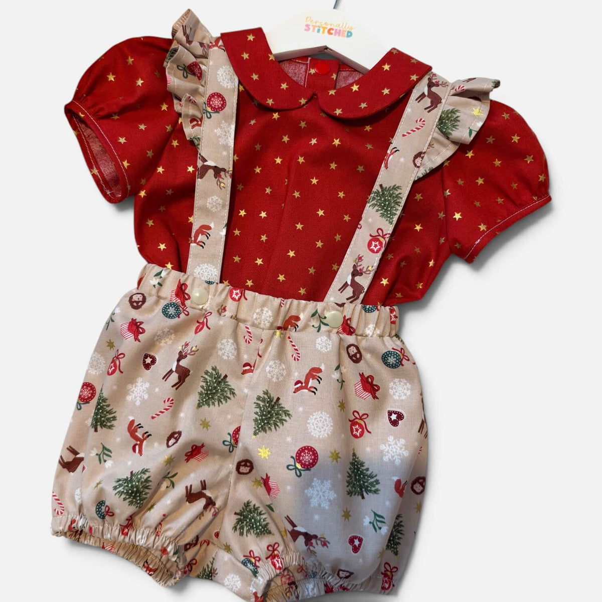 A baby girl's Christmas-themed two-piece romper set featuring a red short-sleeve top with gold stars and ruffled collar, paired with beige suspender shorts covered in a festive holiday print of Santa Claus, trees, and snowflakes.