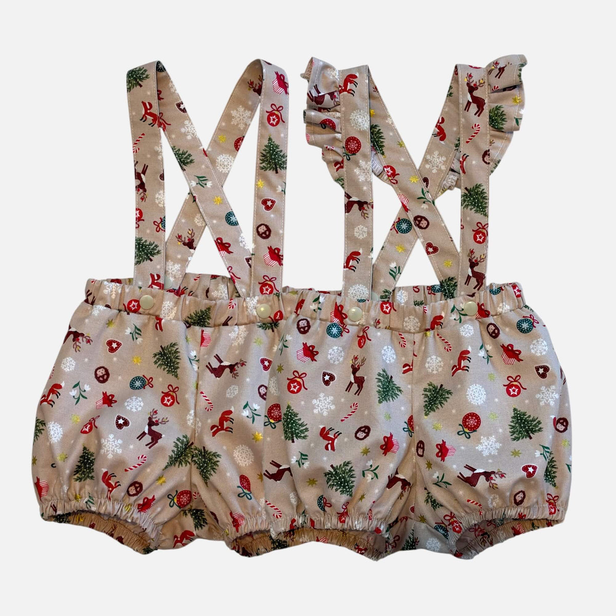 A pair of toddler unisex bloomer suspenders with a Christmas-themed print on a beige fabric, featuring red Santa Clauses, green trees, and white snowflakes, shown against a plain light gray background.