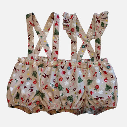 A pair of toddler unisex bloomer suspenders with a Christmas-themed print on a beige fabric, featuring red Santa Clauses, green trees, and white snowflakes, shown against a plain light gray background.