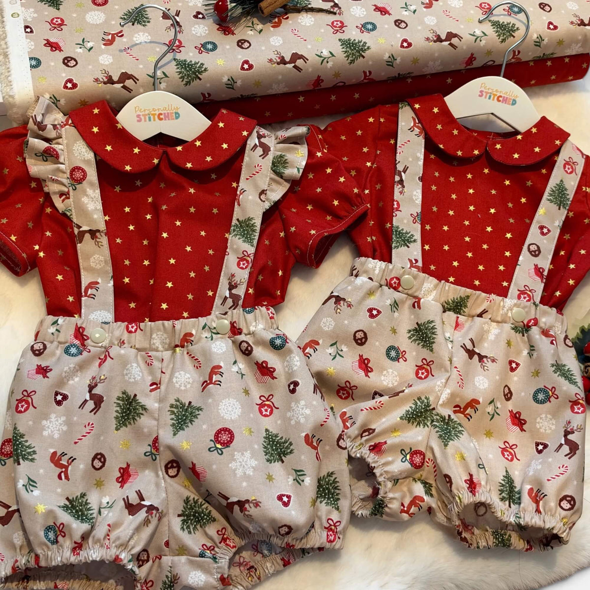 Two baby romper outfits displayed side by side on wooden hangers, featuring red short-sleeve tops with white star patterns and beige bib-style bottoms with a Christmas print. A matching rolled fabric roll is visible in the background.