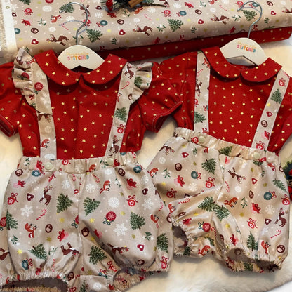 Two baby romper outfits displayed side by side on wooden hangers, featuring red short-sleeve tops with white star patterns and beige bib-style bottoms with a Christmas print. A matching rolled fabric roll is visible in the background.