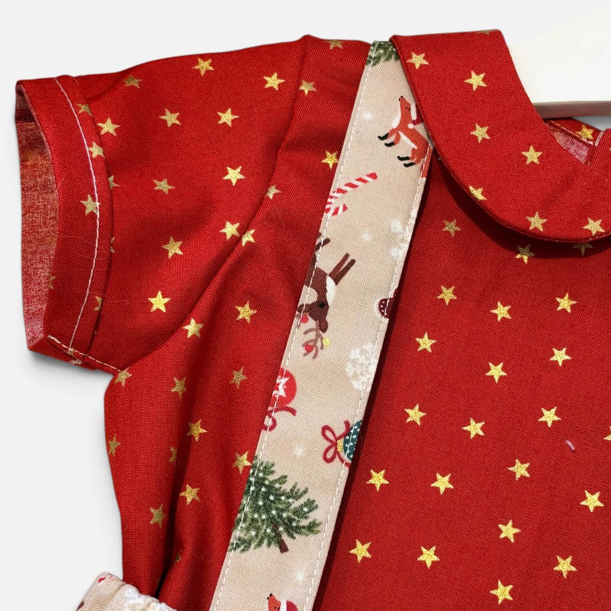 A close-up of a red fabric garment with gold star print, a Peter Pan collar, and a decorative cream ribbon with Christmas illustrations, photographed on a white background.