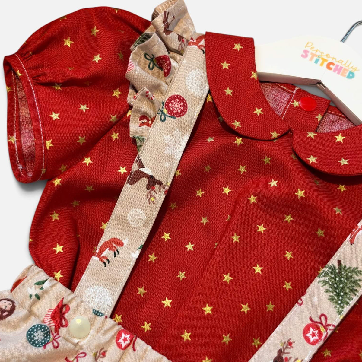 A close-up of a baby outfit set featuring a red short-sleeve top with gold star prints, beige ruffled trim, and matching beige patterned suspenders with holiday motifs, shown on a white hanger against a white background.