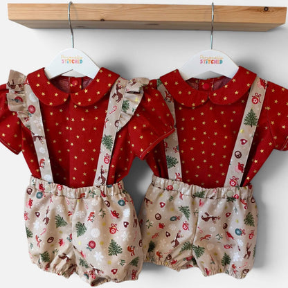 Two baby romper suits hanging on white hangers from a wooden shelf. The set features a red short-sleeve top with gold star patterns and beige suspenders with a Christmas print of trees, ornaments, and reindeer.