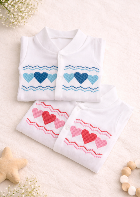 White baby sleep suits with embroidered heart designs in pink and blue, neatly folded on a soft cream background, suitable for Valentine’s Day babywear.