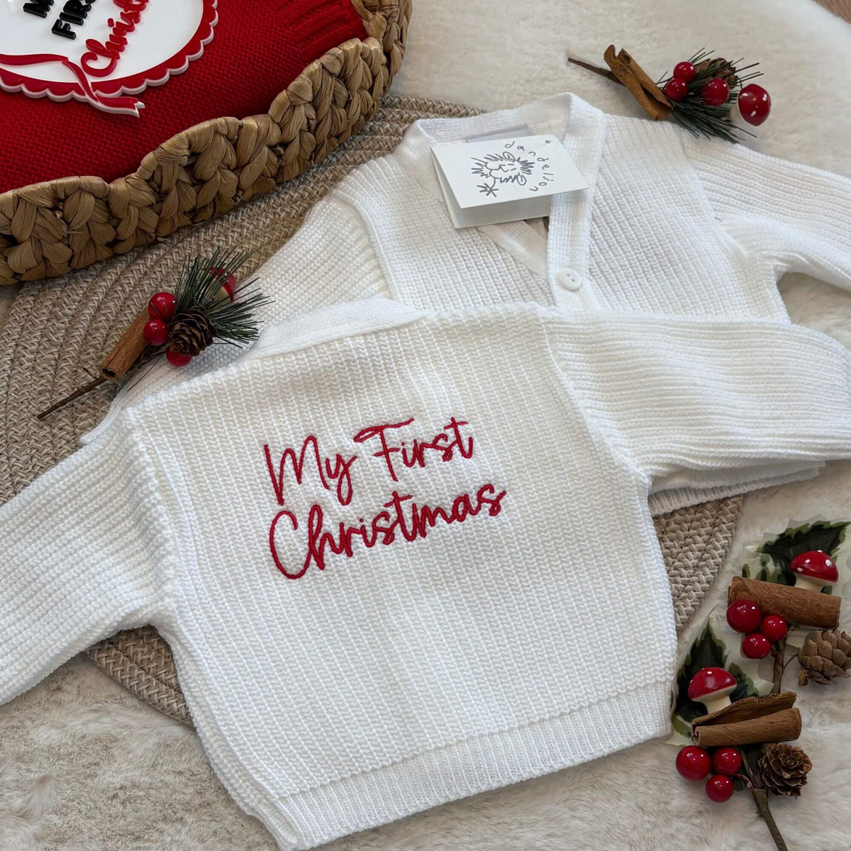 A white ribbed knit baby cardigan laid flat on a beige rug with holiday decorations. The cardigan has 'My First Christmas' embroidered in red script on the back and a white paper tag attached at the collar.