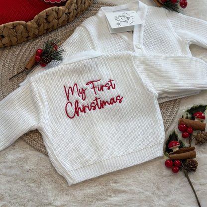 A white ribbed knit baby cardigan laid flat on a textured surface, with red embroidered text reading 'My First Christmas' on the back. A small branded tag is attached near the collar, and the cardigan is styled with holiday props including berries, pinecones, cinnamon sticks, and woven mats.