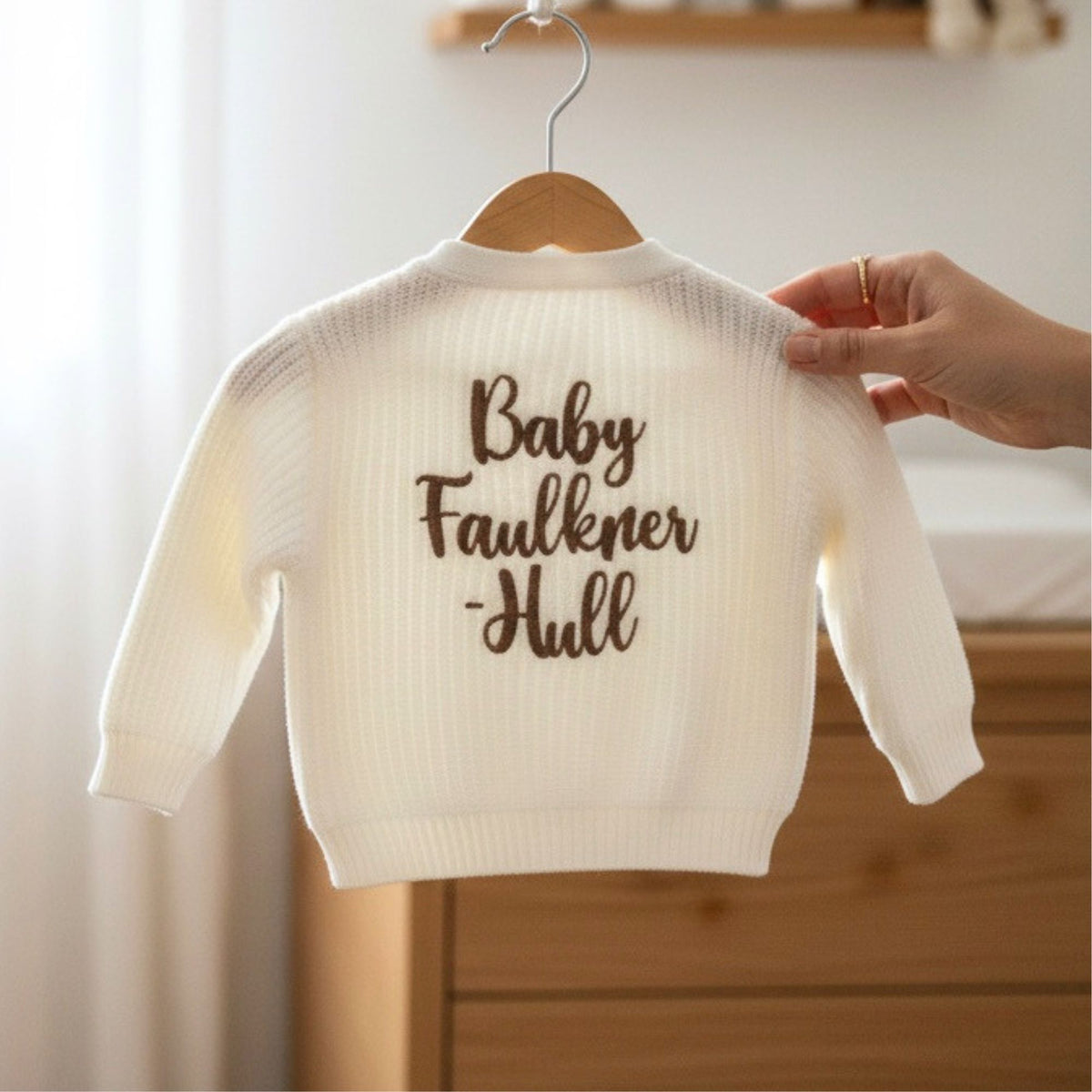 A white knit baby cardigan with babies surname embroidered on it in a light brown thread.