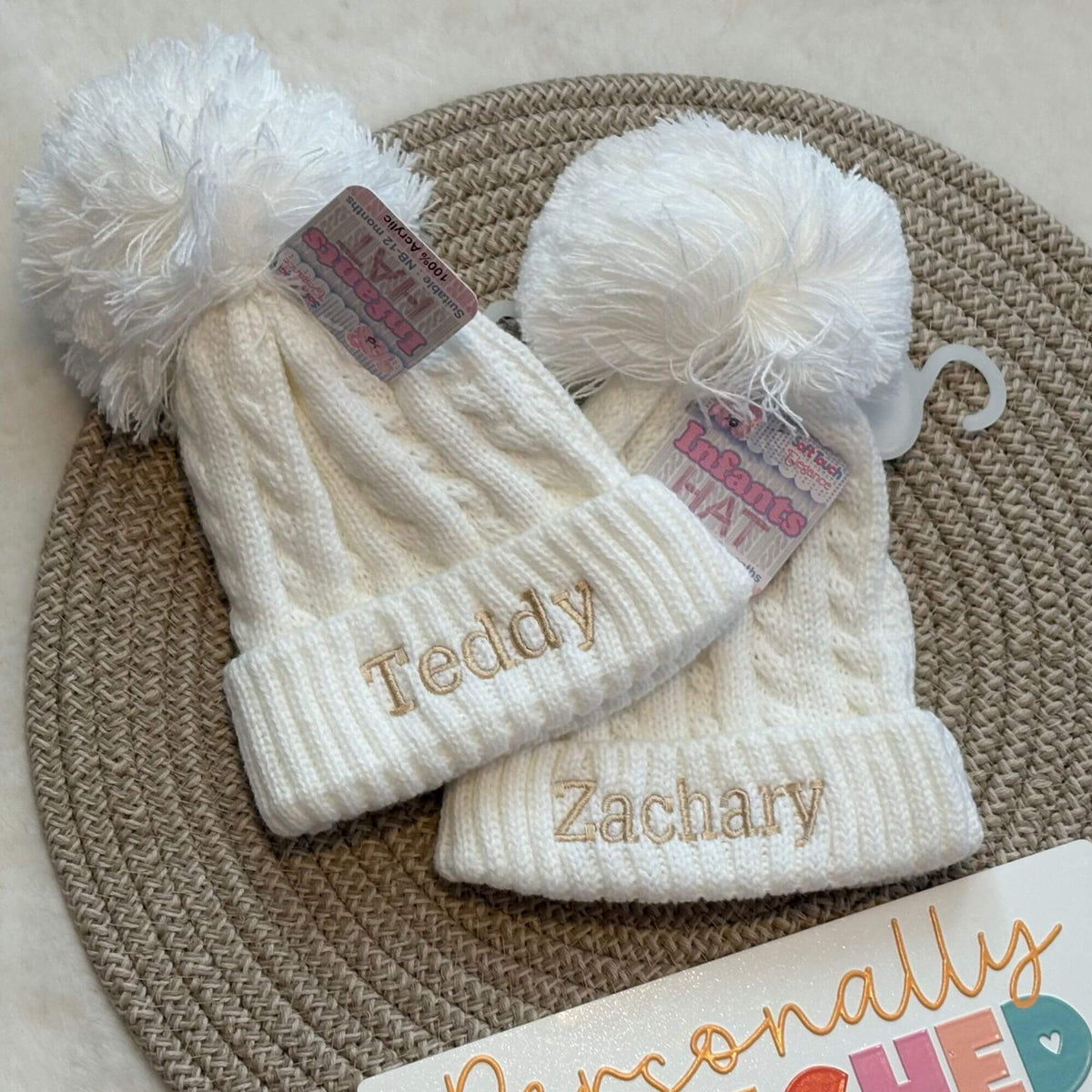 Two personalised white cable-knit baby beanies with white faux-fur pom-poms, laid flat on a woven round mat. Hats are personalised using embroidery in a beige thread 